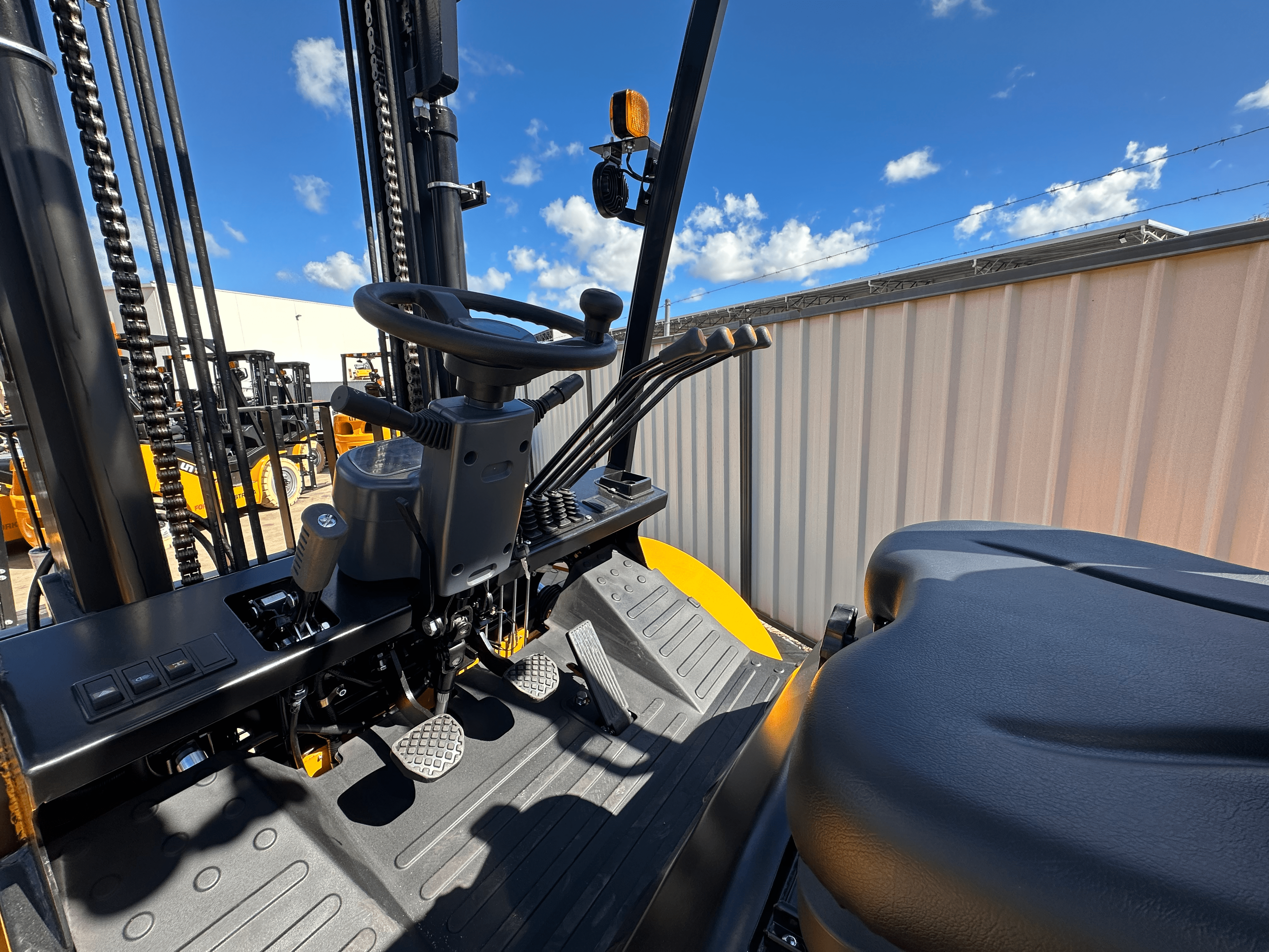 The driver's seat and controls of a forklift. The controls include a steering wheel, pedals, levers, and buttons. The seat is black and the background is a blue sky with some clouds.