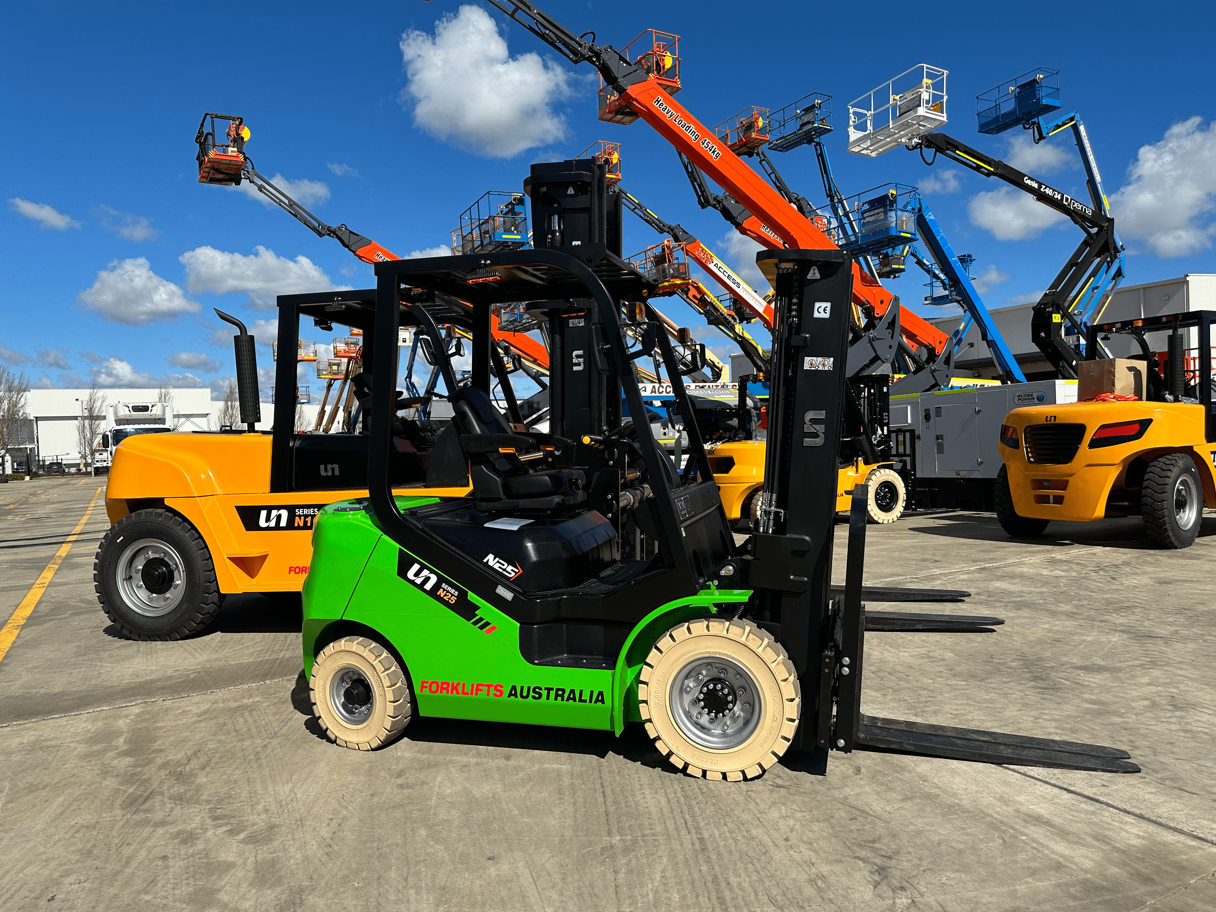 A green forklift with the brand name "UN" and the label "FORKLIFTS AUSTRALIA" on the side. It is parked in a yard with several other yellow forklifts and aerial work platforms in the background. The sky is blue with some clouds.