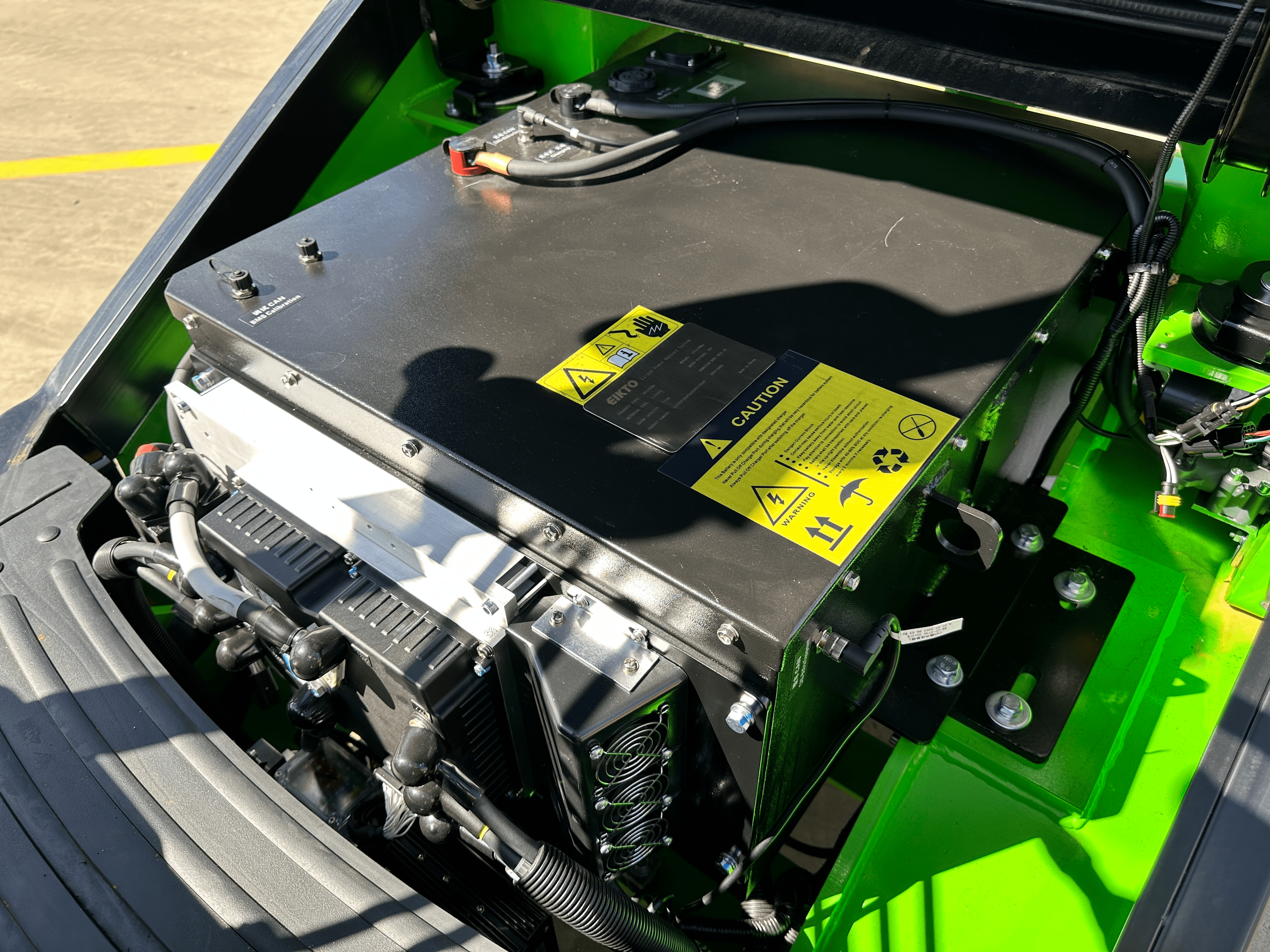 A close-up of the battery compartment of a green electric forklift. The battery is covered by a black panel with warning signs and labels. There are also cooling fans and wires visible inside the compartment.