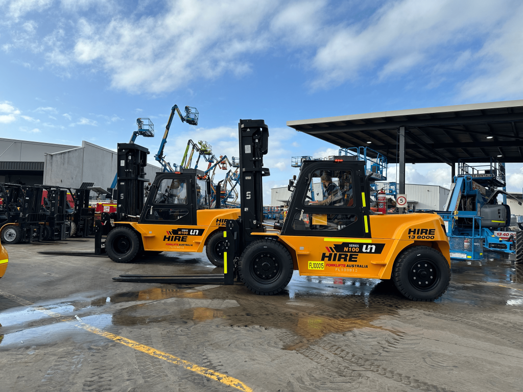 Several yellow forklifts are parked in a row in a large outdoor area. The forklifts have the brand name "UN" and the word "HIRE" written on them. There are other pieces of heavy machinery visible in the background.