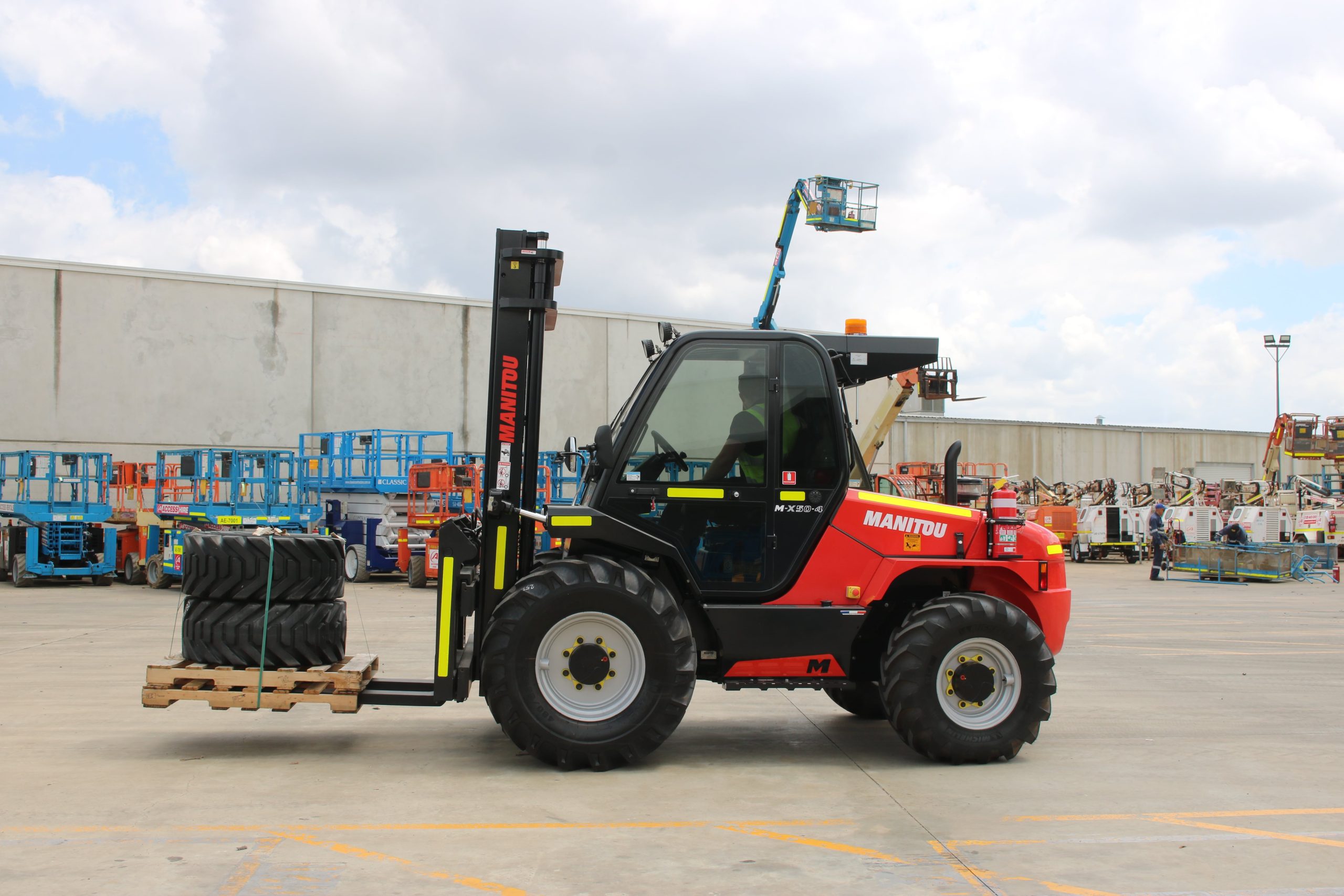 A red forklift with the brand name "MANITOU" on the side. It has a black cab, black tires, and a raised mast with a basket attachment. The forklift is lifting a pallet of tires in a large outdoor area. There are other pieces of heavy machinery visible in the background.