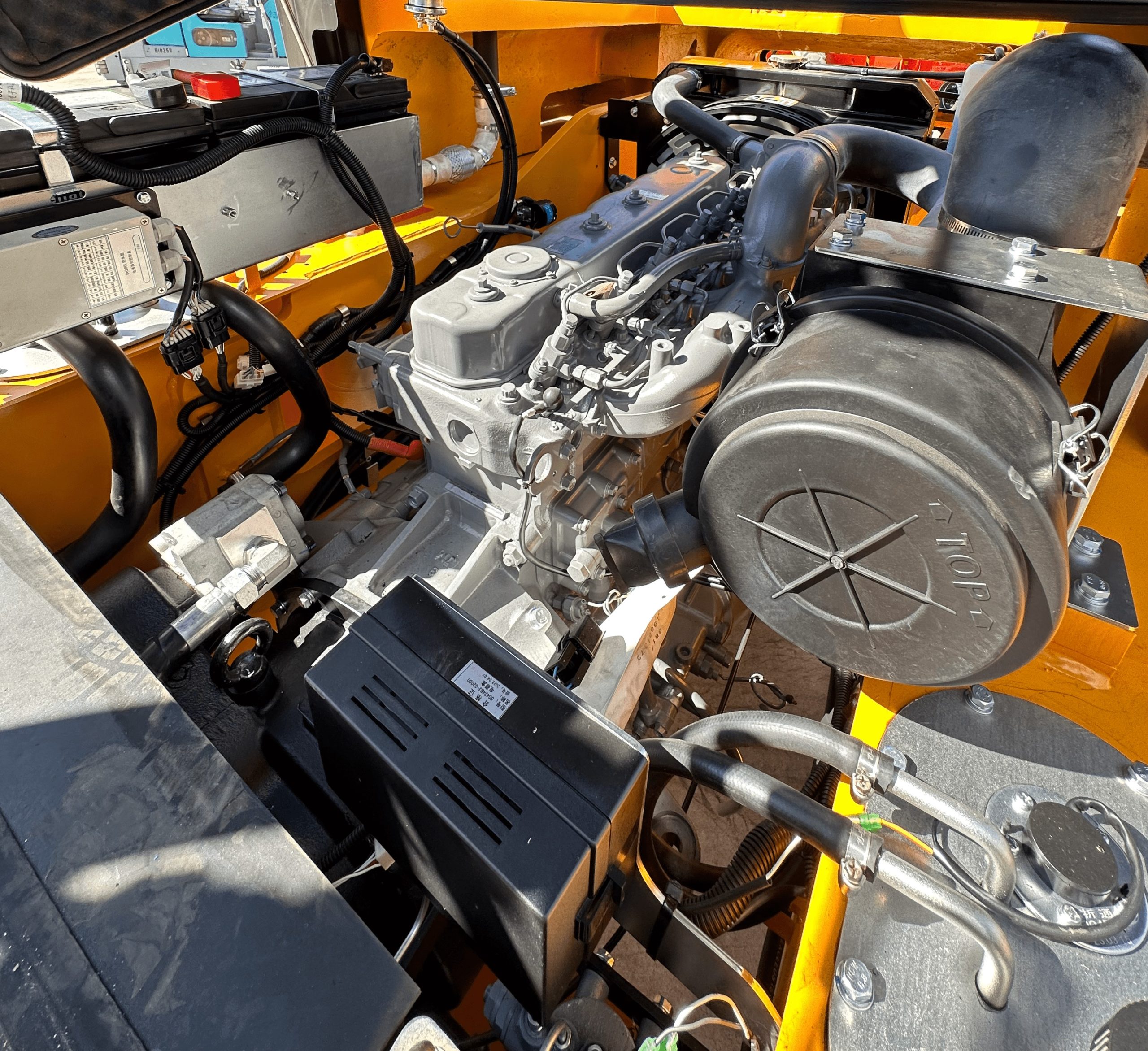 A close-up view of a vehicle's engine compartment, showing various components like the engine, air filter, electrical connections, and hoses. The area is well-lit, and some parts are marked with labels and numbers. The surrounding structure is painted yellow.