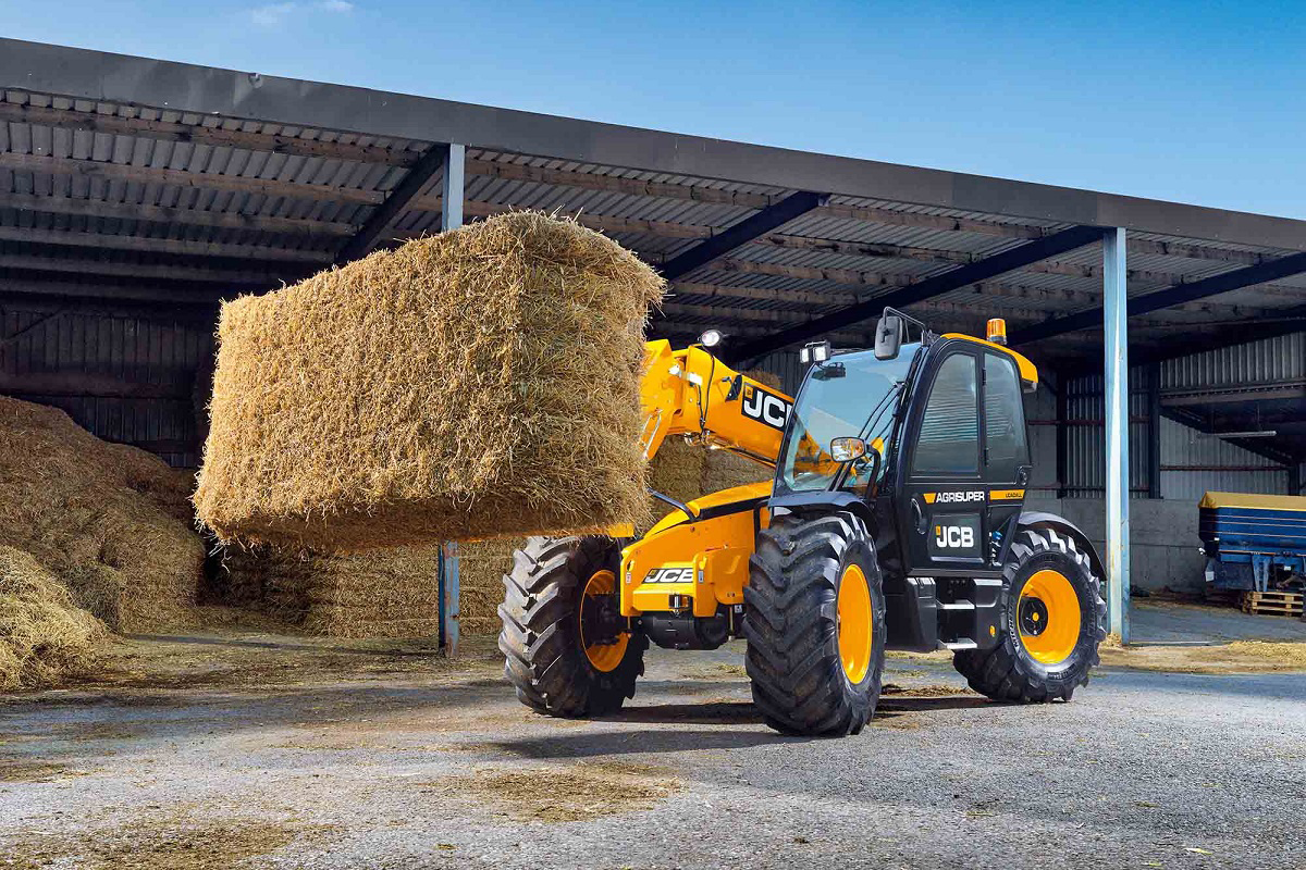A yellow JCB telehandler lifting a large bale of hay in front of a covered storage area. The background features a clear blue sky and a partially visible machinery shed. The scene is set in a farmyard.