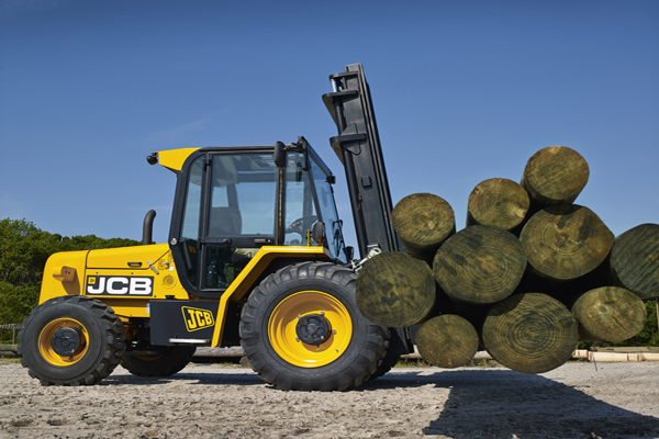 A yellow JCB forklift is lifting a bundle of large, round wooden logs in a sunlit, outdoor setting. The machine is positioned on a gravel surface, with green trees visible in the background.