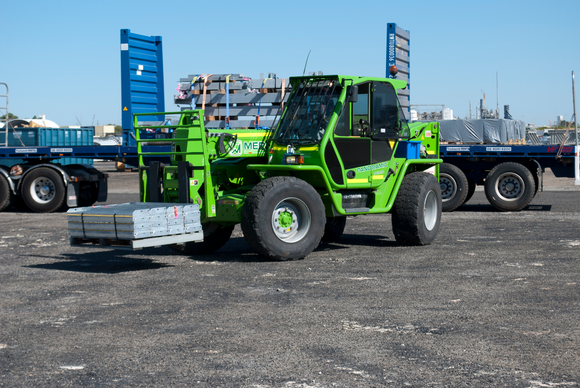 A green telescopic forklift, also known as a telehandler, lifts a pallet of materials in an outdoor industrial area. Two flatbed semi-trailers loaded with cargo are visible in the background. The ground is paved, and the sky is clear.