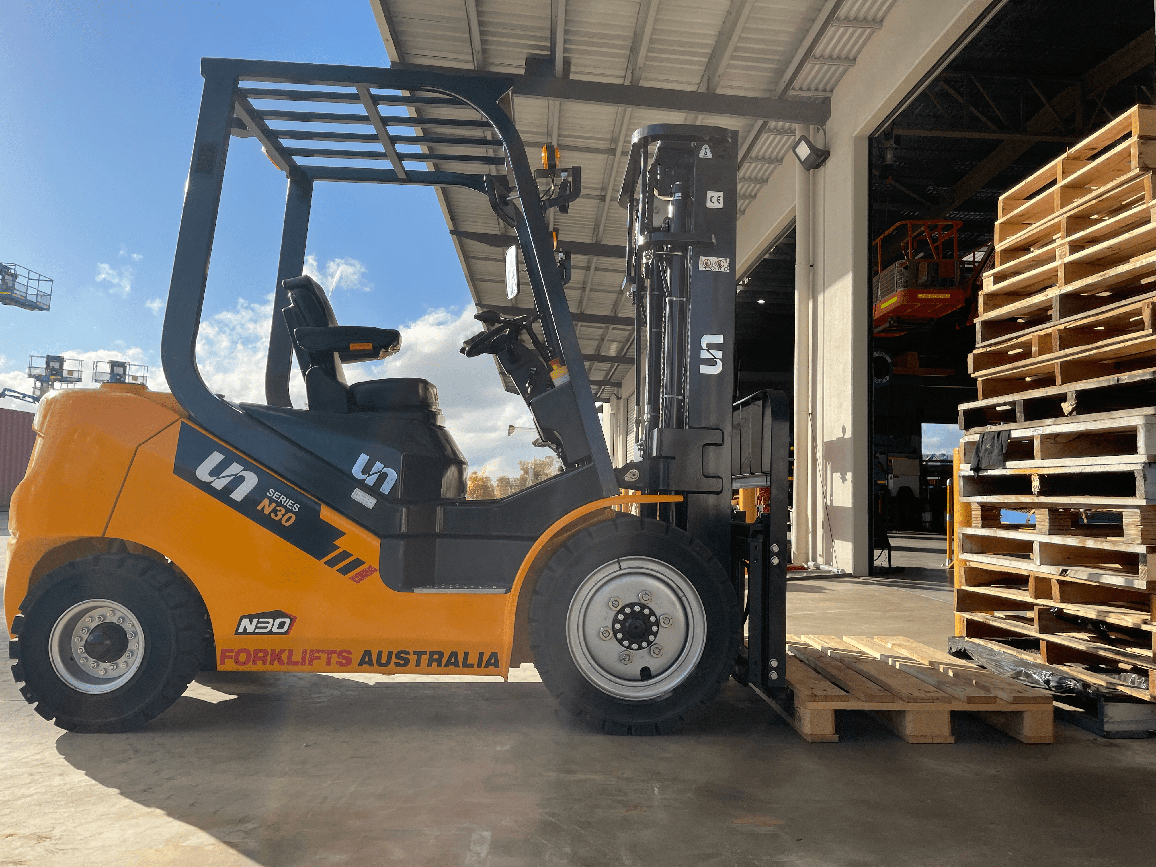 A bright yellow forklift truck sits idly in front of an industrial warehouse. The forks are positioned to lift wooden pallets stacked nearby. The warehouse interior is slightly visible, showing more equipment and stacked items. Sunlight casts shadows on the ground.