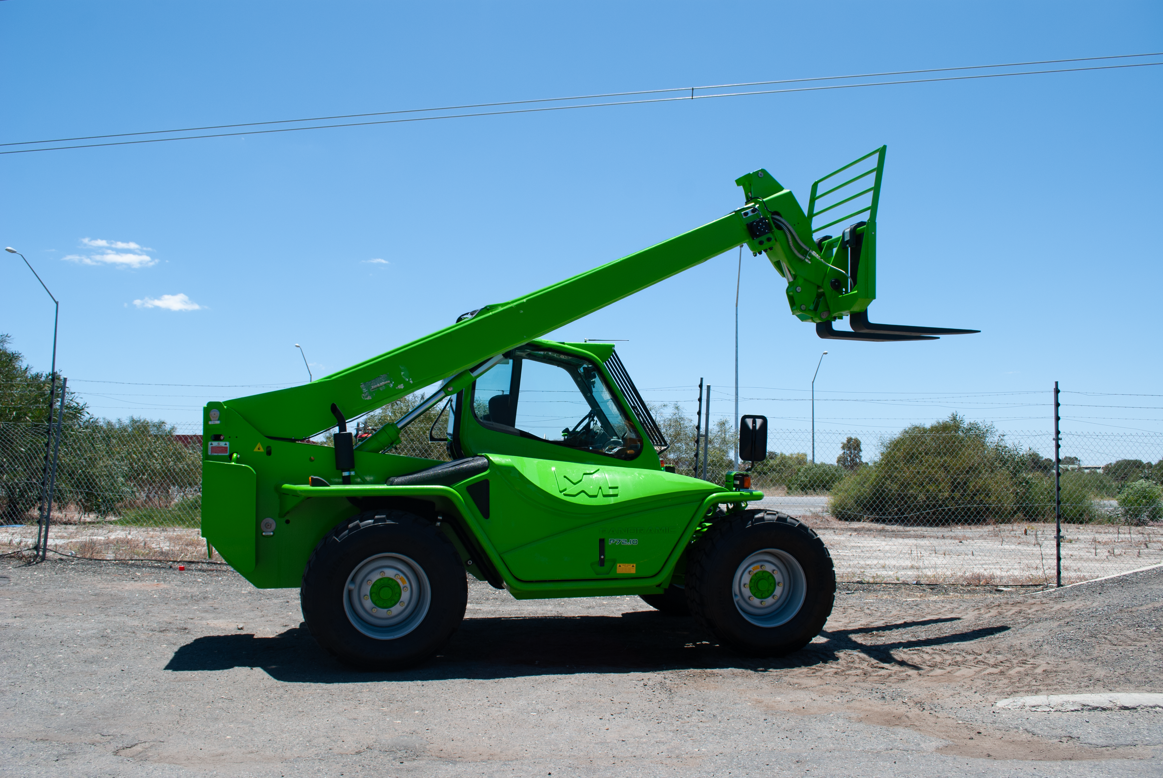 A green telescopic handler forklift is lifting a load of stacked materials on a flat surface in an industrial area. Several trailers, one with additional stacked materials, are visible in the background against a clear blue sky.
